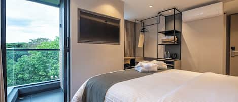 A modern hotel room with a large bed, a wardrobe, a seating area, and a view of greenery outside.