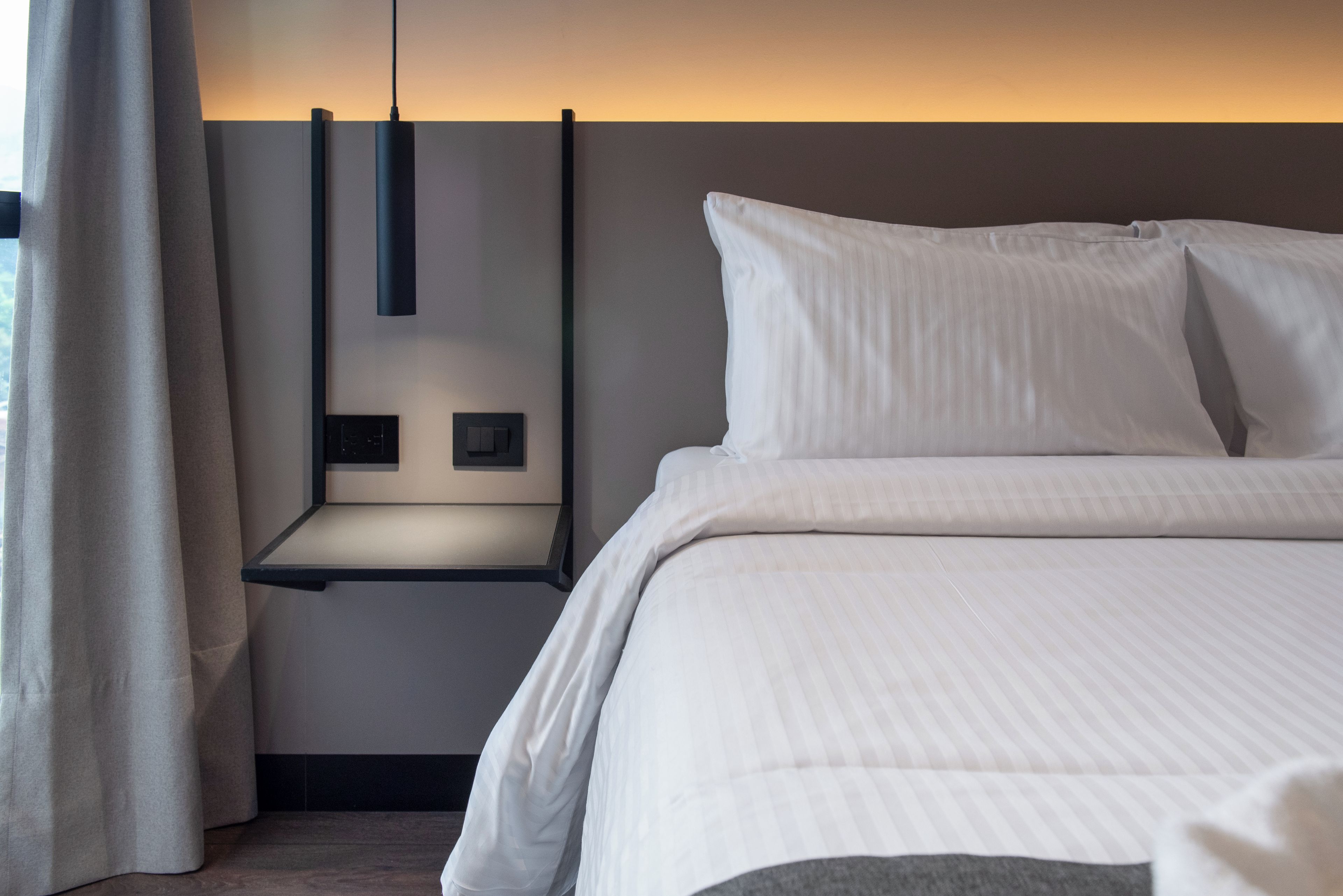Habitación de hotel moderna con una cama bien tendida, una mesita de noche con lámpara y un tablero de interruptores en la pared.