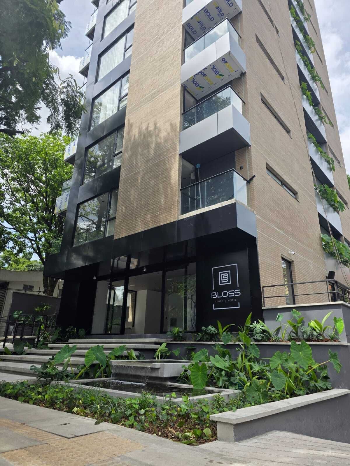 Un hôtel moderne, avec un logo bien visible, de grandes fenêtres et un espace d’entrée aménagé avec des éléments paysagers.