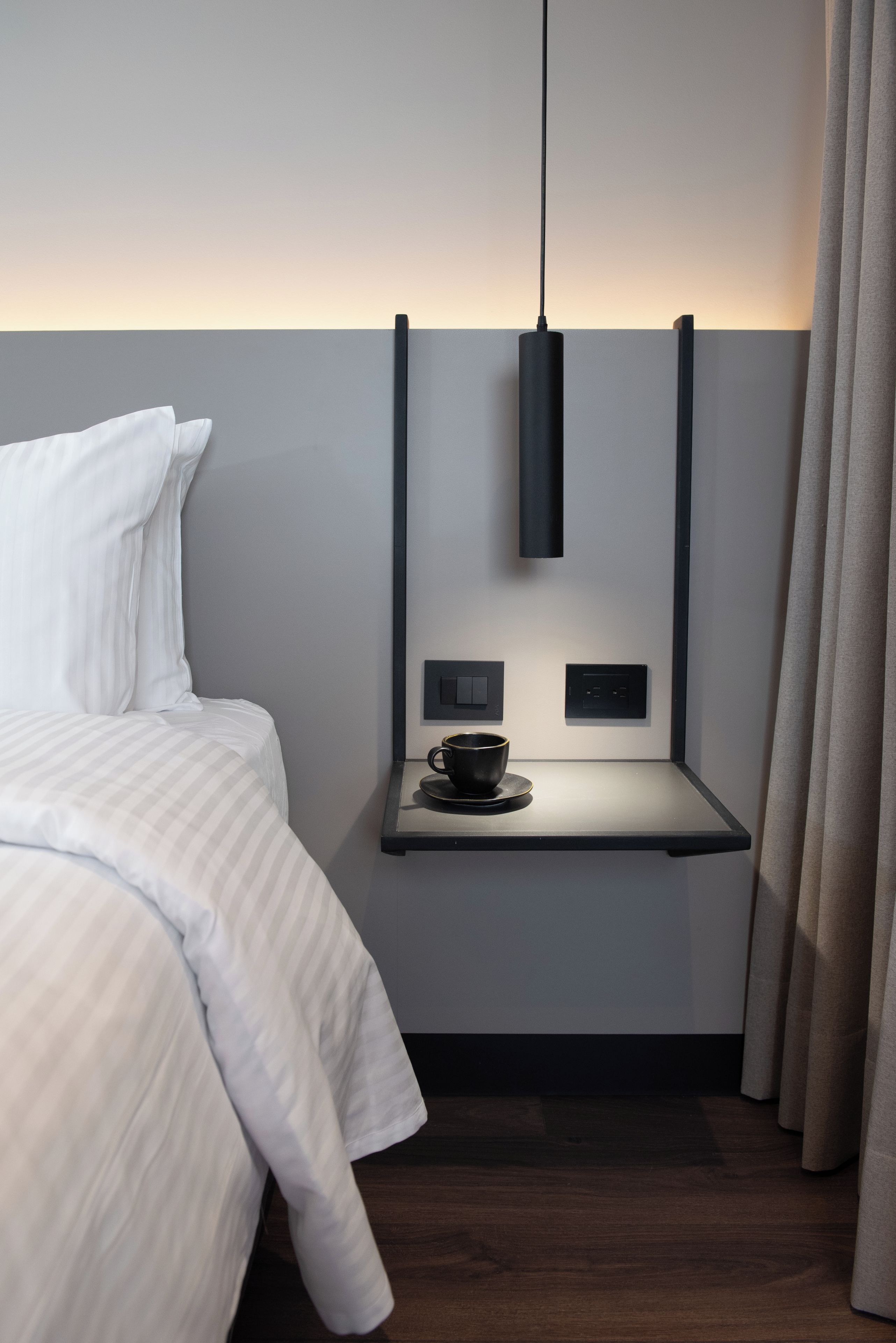 A modern hotel room with a bed, a bedside table with a cup, and a pendant light.