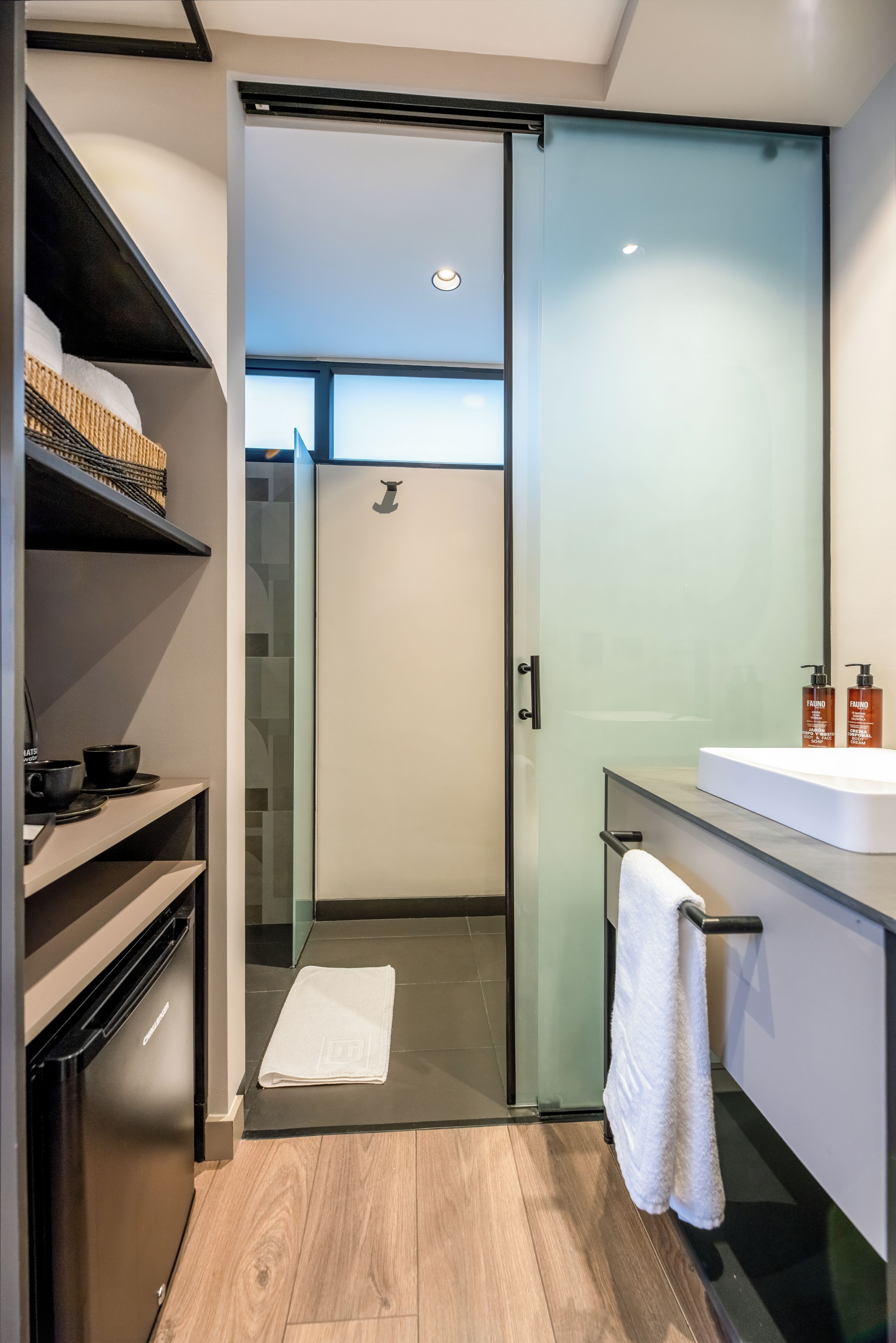 Une salle de bain moderne avec une porte de douche en verre, un lavabo blanc avec une serviette et une étagère contenant des articles de toilette.