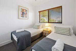 Een hotelkamer met twee bedden, een nachtkastje met een lamp, een raam met zonwering en een lijst met een afbeelding aan de muur.