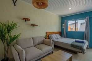 A room with a sofa, a bed, a small table, a hanging light fixture, and a window with curtains. - Aruba Vibe Apartments 8 | Charming Studio Near the Beaches and Downtown (Oranjestad)