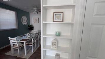 A dining area with a table set for four, white chairs, a ceiling fan, and a wall with framed pictures and a shelf with decorative items.