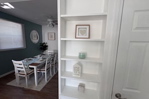 Un coin repas avec une table dressée pour quatre personnes, des chaises blanches, un ventilateur de plafond et un mur orné de tableaux et d’une étagère où sont disposés des objets décoratifs.