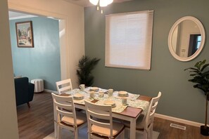 A dining area with a table set for four, white chairs, a round mirror, and a window with blinds. - The Gem On The Block (Springfield)