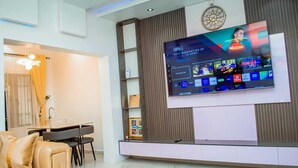 A modern living room with a flat-screen TV mounted on the wall, a dining area with a table and chairs, and a sofa. - The Haven | Modern 2BR En-suite Flat (Ibadan)