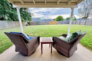 An outdoor patio with wicker furniture and a table, surrounded by a wooden fence and a grassy yard.