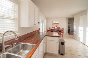 Una cocina con encimera de madera, pileta de acero inoxidable, gabinetes blancos y un comedor con mesa y sillas.