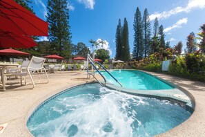 Un jacuzzi con piscina, rodeado de sillones de descanso y sombrillas, con árboles altos y un cielo despejado al fondo.