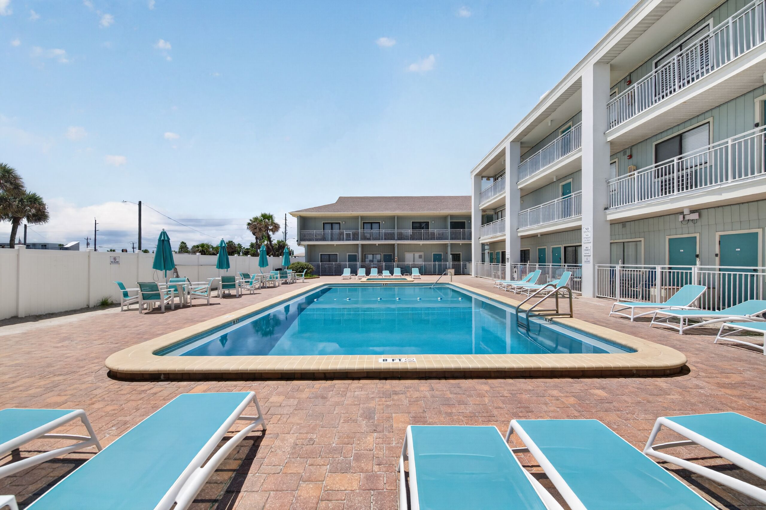 A swimming pool with lounge chairs and umbrellas, a multi-story building with balconies, and a clear sky.