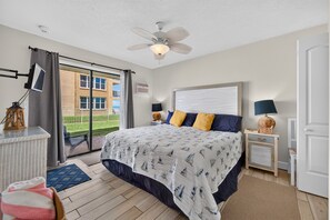 Un dormitorio con una cama grande, ventilador de techo, ventana con cortinas y vista a un edificio.