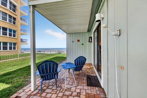 A covered outdoor seating area with two chairs and a table, a white door with a doormat, and a view of the ocean. - Sun and Sand Getaway @ Coastal Waters (New Smyrna Beach)