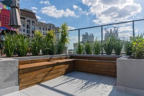 A rooftop terrace with a wooden bench, potted plants, and a view of city buildings.
