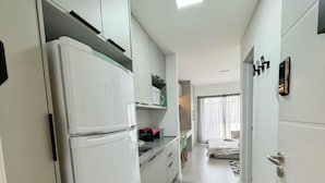 Une cuisine compacte avec des meubles blancs, un réfrigérateur et un four à micro-ondes. On y trouve un petit coin repas avec des chaises et une table. La pièce présente une palette de couleurs neutres et un design moderne.