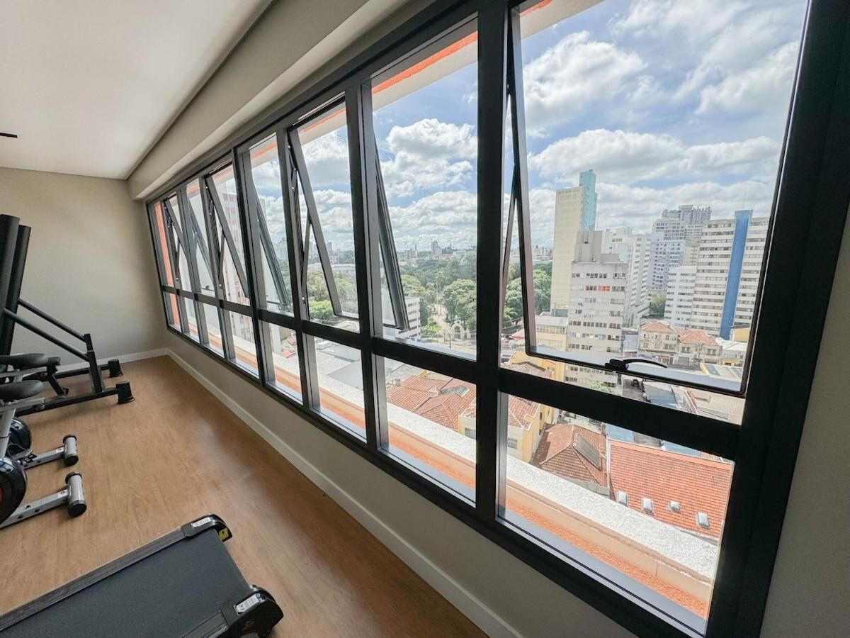 Ginásio com equipamentos de exercício e amplas janelas com vista para a paisagem urbana.