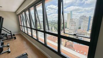 Ginásio com equipamentos de exercício e amplas janelas com vista para a paisagem urbana.
