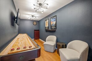 Ein Zimmer mit Shuffleboard-Tisch, zwei Stühlen, Dartscheibe und Wandbildern.