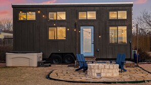 A modern, two-story building with a flat roof, multiple windows, and a blue door. There is a stone fire pit with two blue chairs and a small table in the front yard. - Henley, Tiny Home Escape with Hot Tub & Fire Pit in Fredericksburg (Fredericksburg)