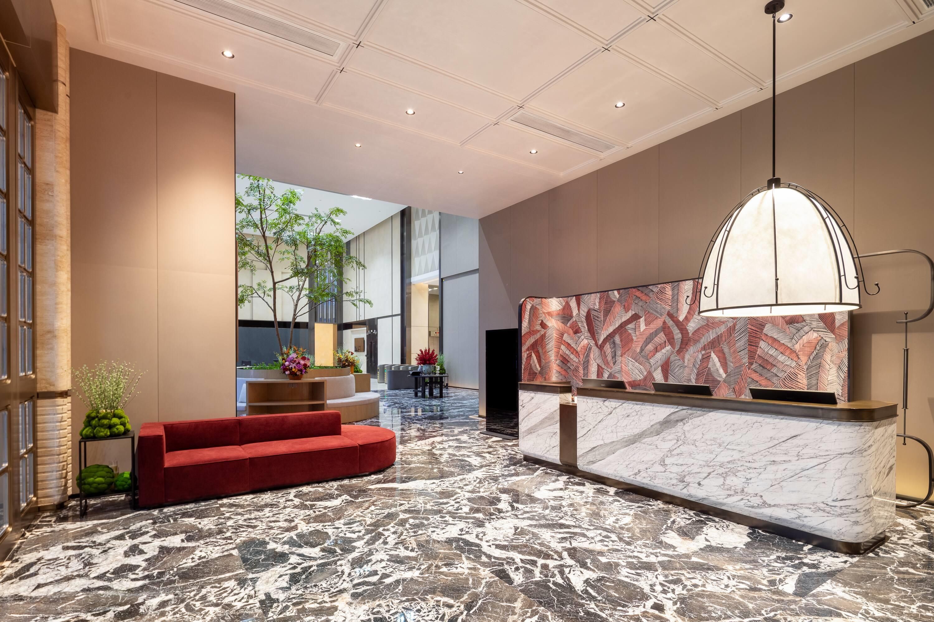 A hotel lobby with a reception desk, a red sofa, a marble floor, and a large window overlooking greenery.
