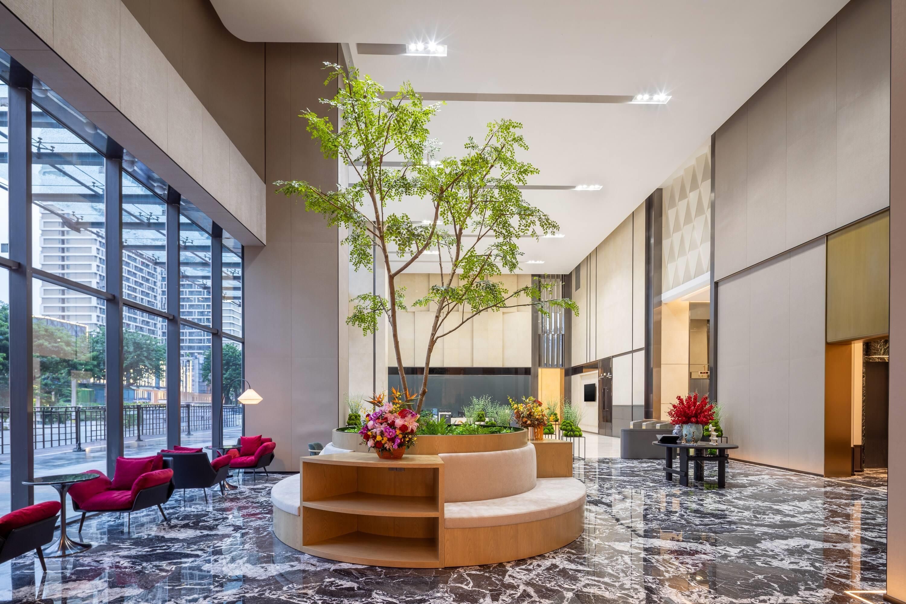 A spacious lobby with a central wooden planter, seating area, and large windows offering views of the city.