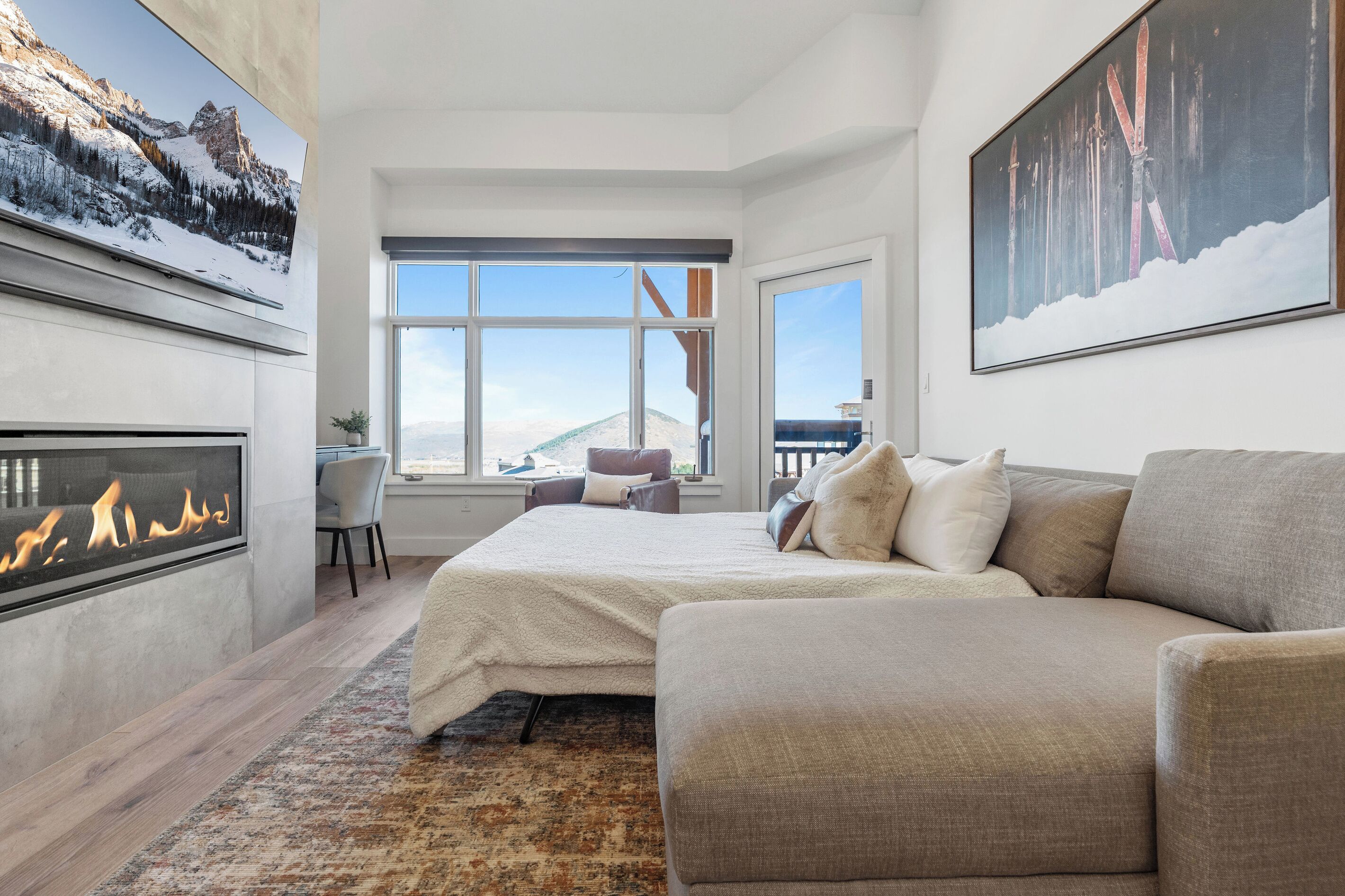 A modern bedroom with a fireplace, a large bed, a sofa, and a view of mountains through the windows.