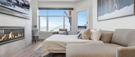 Un dormitorio moderno con chimenea, una cama grande, un sofá y vistas a las montañas a través de las ventanas.