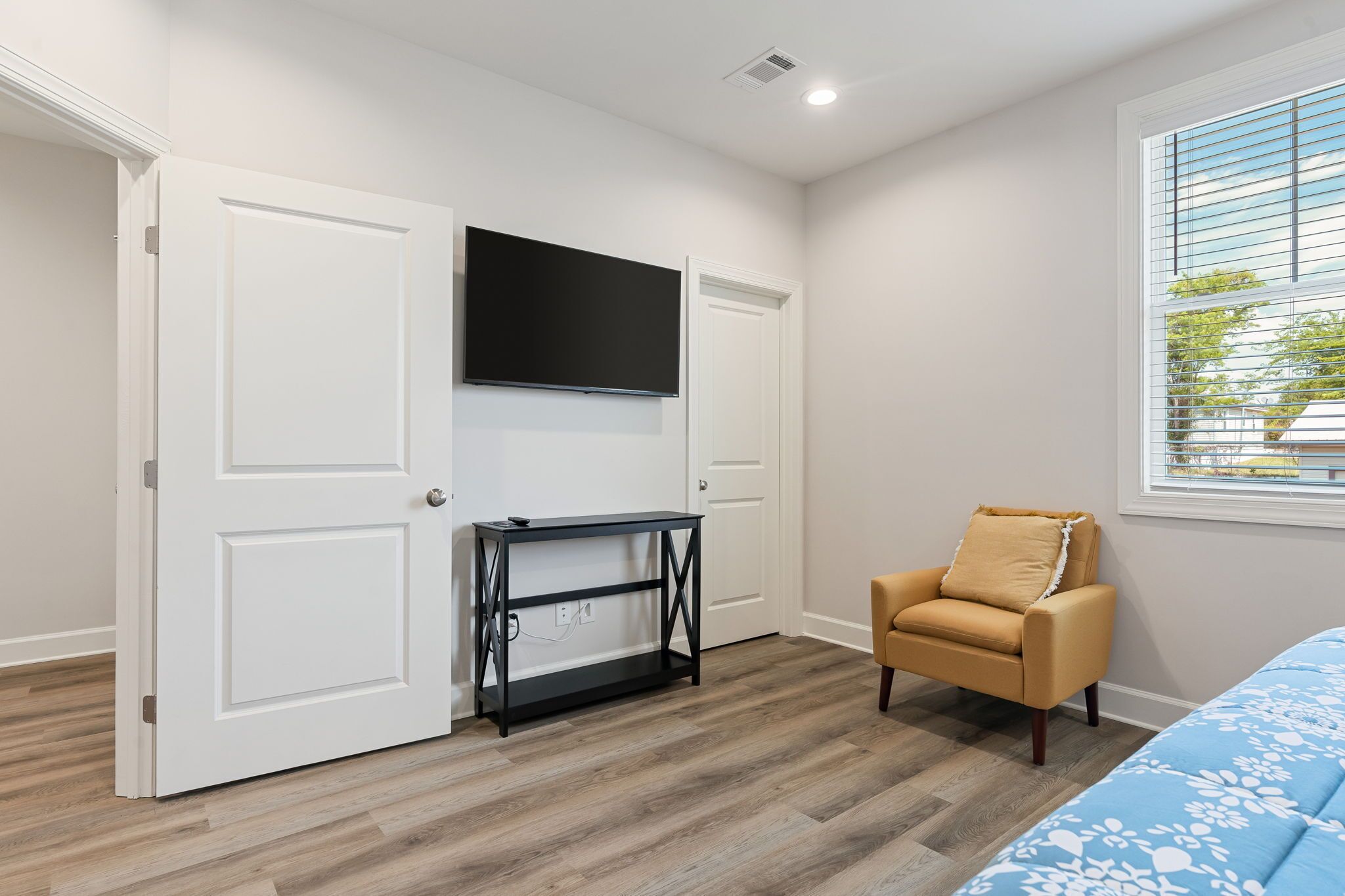 A bedroom with a TV mounted above a black console table, a beige armchair, and a bed with a blue floral bedspread.