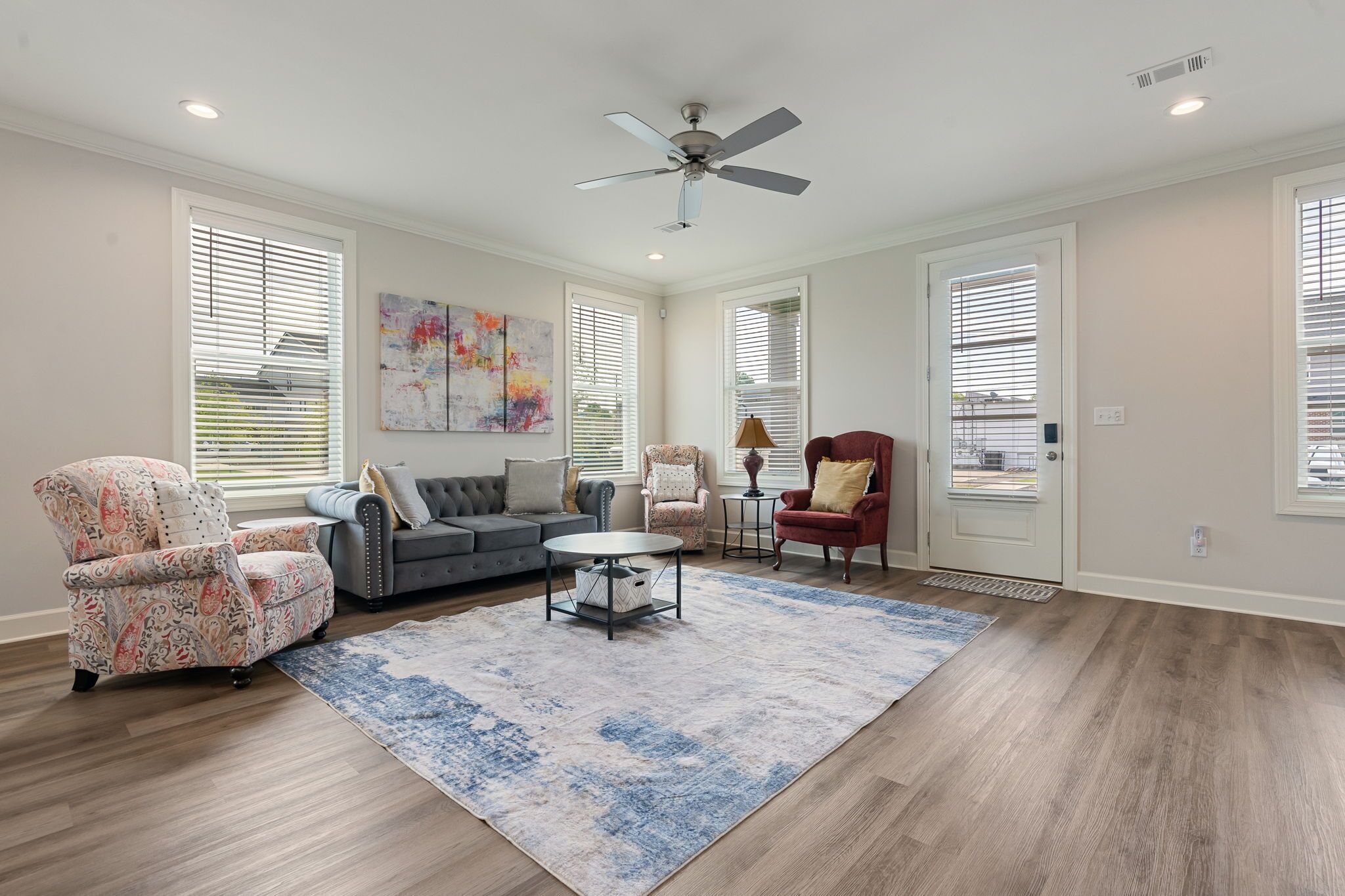 A spacious living room with hardwood flooring, a ceiling fan, a gray sofa, a patterned armchair, a red armchair, and a coffee table.