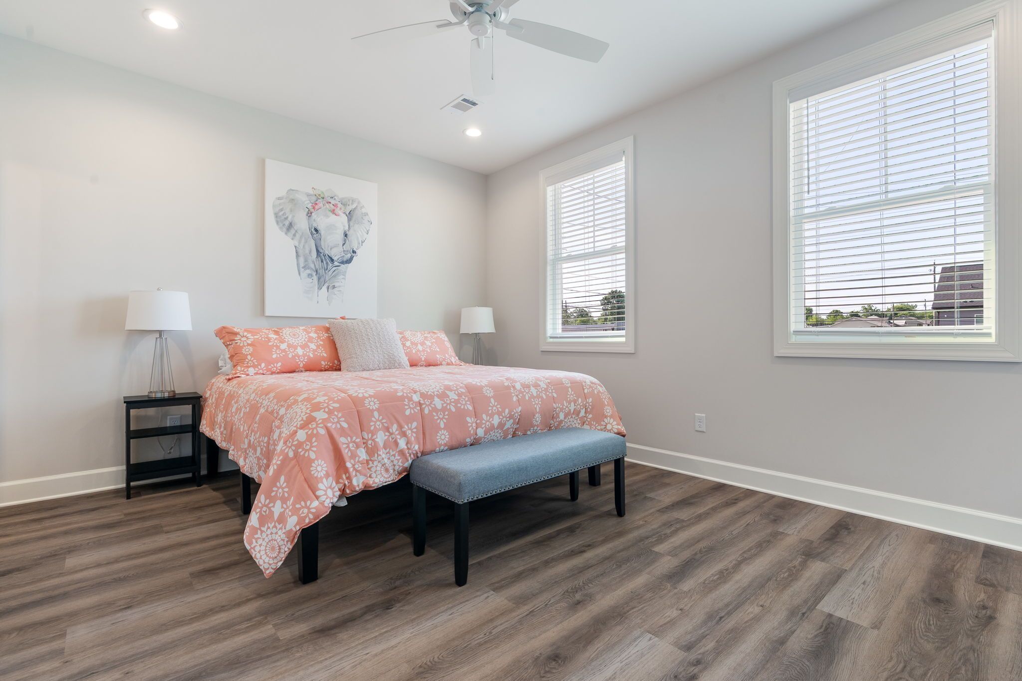 A bedroom with a bed, a bench, two nightstands, a ceiling fan, and a window with blinds.