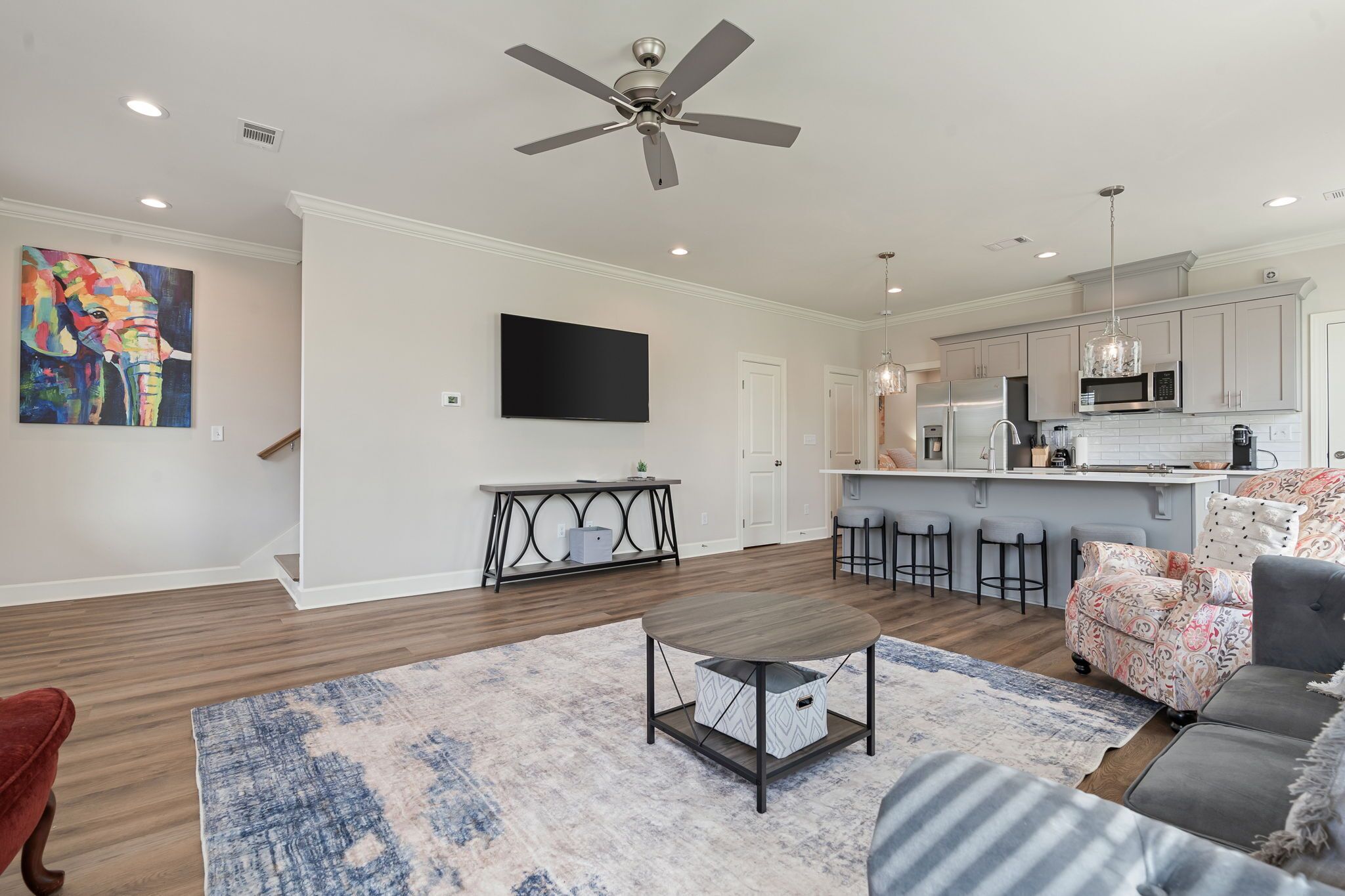 A modern living room with a ceiling fan, a flat-screen TV, and a kitchen area with a microwave and bar stools.