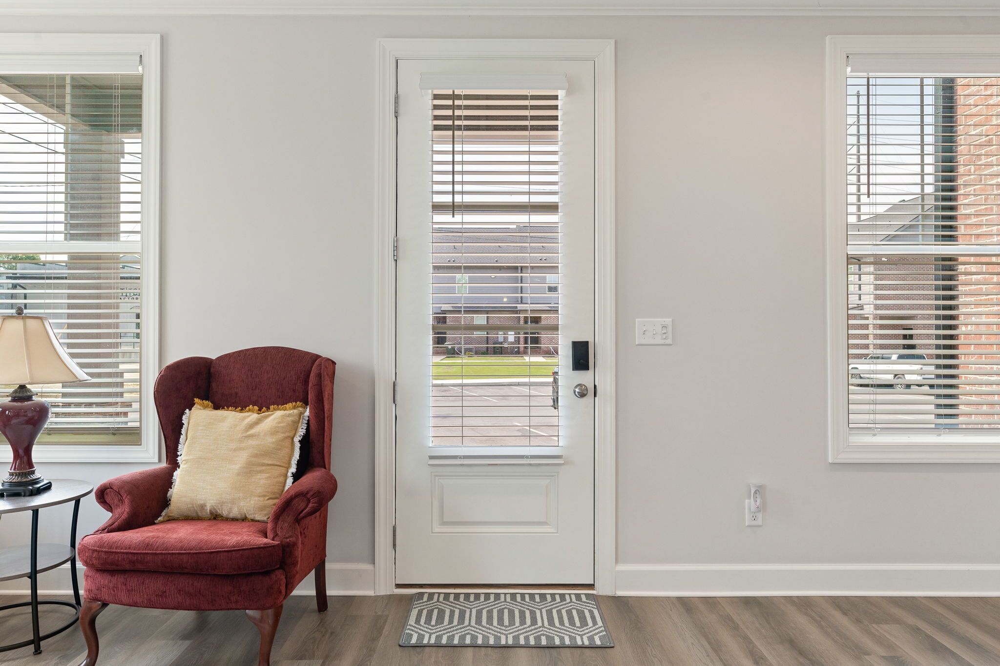 A room with a white door, a red armchair with a cushion, a small table with a lamp, and a patterned rug.