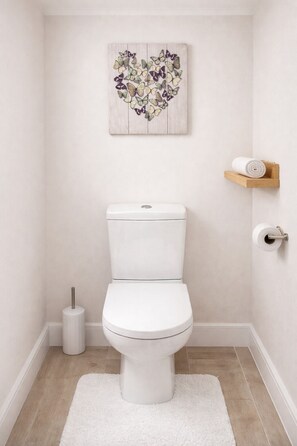 A bathroom with a white toilet, a white rug, a toilet paper holder, and a wall-mounted decorative piece. - Cosy 4 Bed Family Home near Derby City Centre  Rolls Royce (Derby)