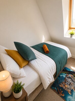 Una cama bien hecha con sábanas blancas, un plaid verde aguamarina y un plaid naranja doblado. Hay una lámpara blanca sobre una mesita de noche, una planta pequeña en maceta y una almohada decorativa con estampado de leopardo.