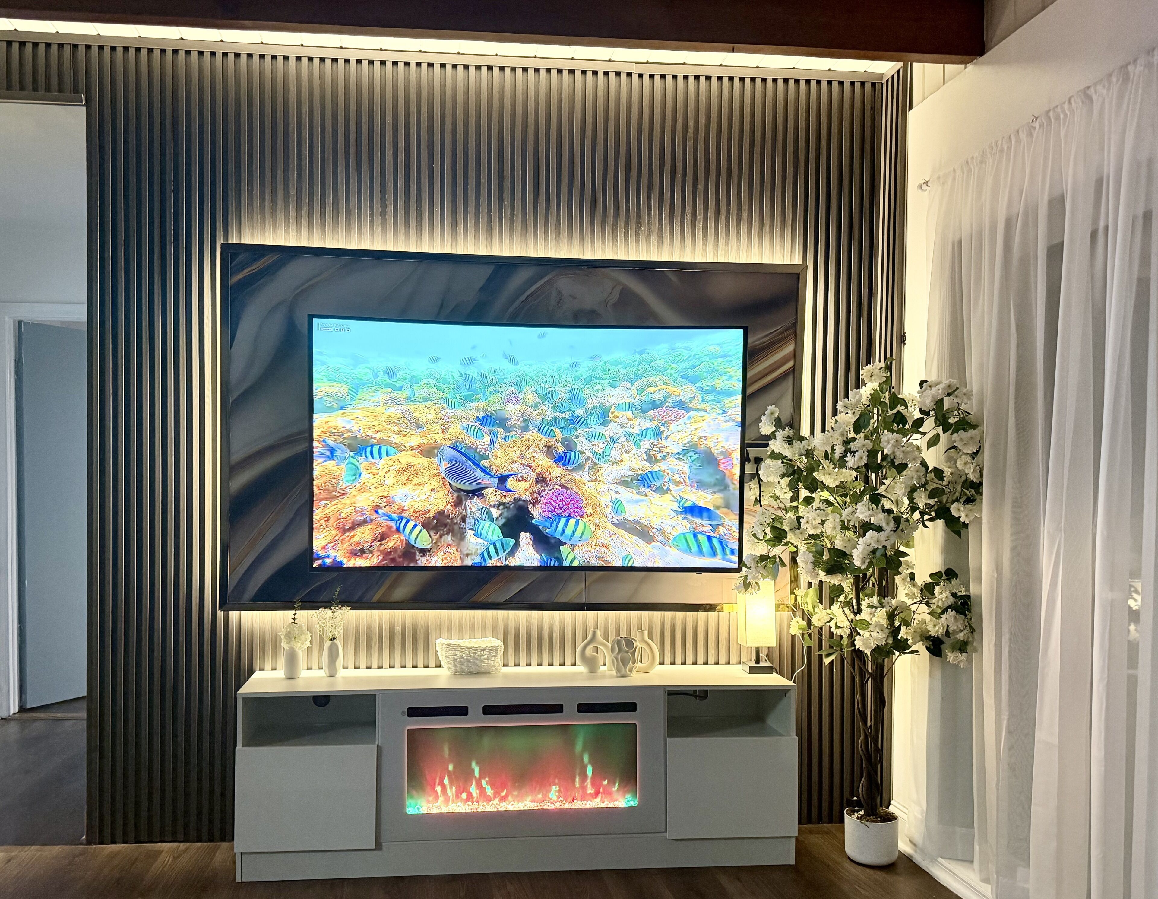 Un téléviseur à écran plat fixé au mur, avec un panneau décoratif derrière, une console blanche en dessous, et une cheminée, ainsi qu’une plante en pot sur la droite.
