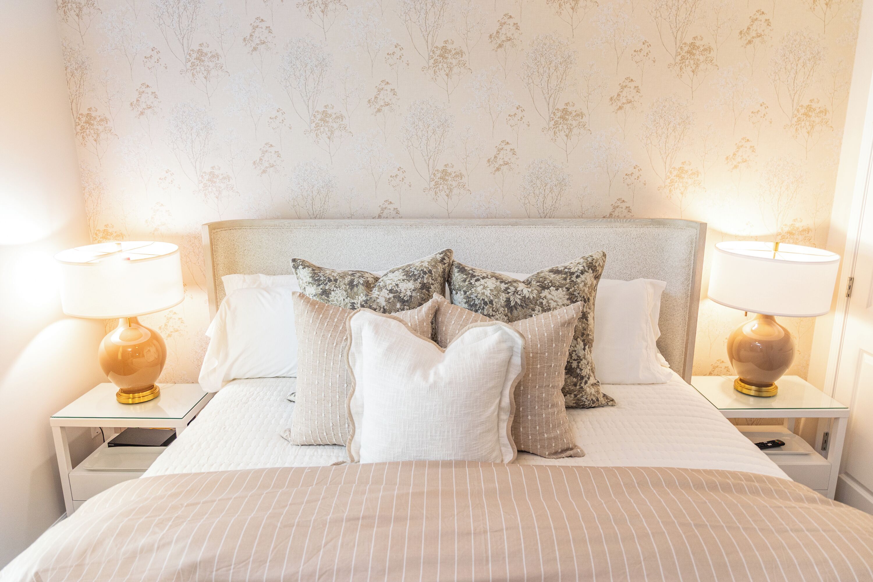 Un lit bien fait, recouvert d’une couvre-lit à rayures, flanqué de deux tables de chevet avec des lampes, le tout adossé à un mur orné d’un motif floral.
