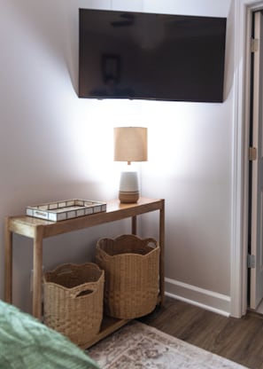 A room with a television mounted on the wall, a table with a lamp and baskets, and a door.