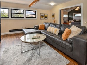 A living room with a leather sofa, a glass coffee table, and a view of the outdoors through large windows. - SunHaven at Stone Ridge (Boulder)