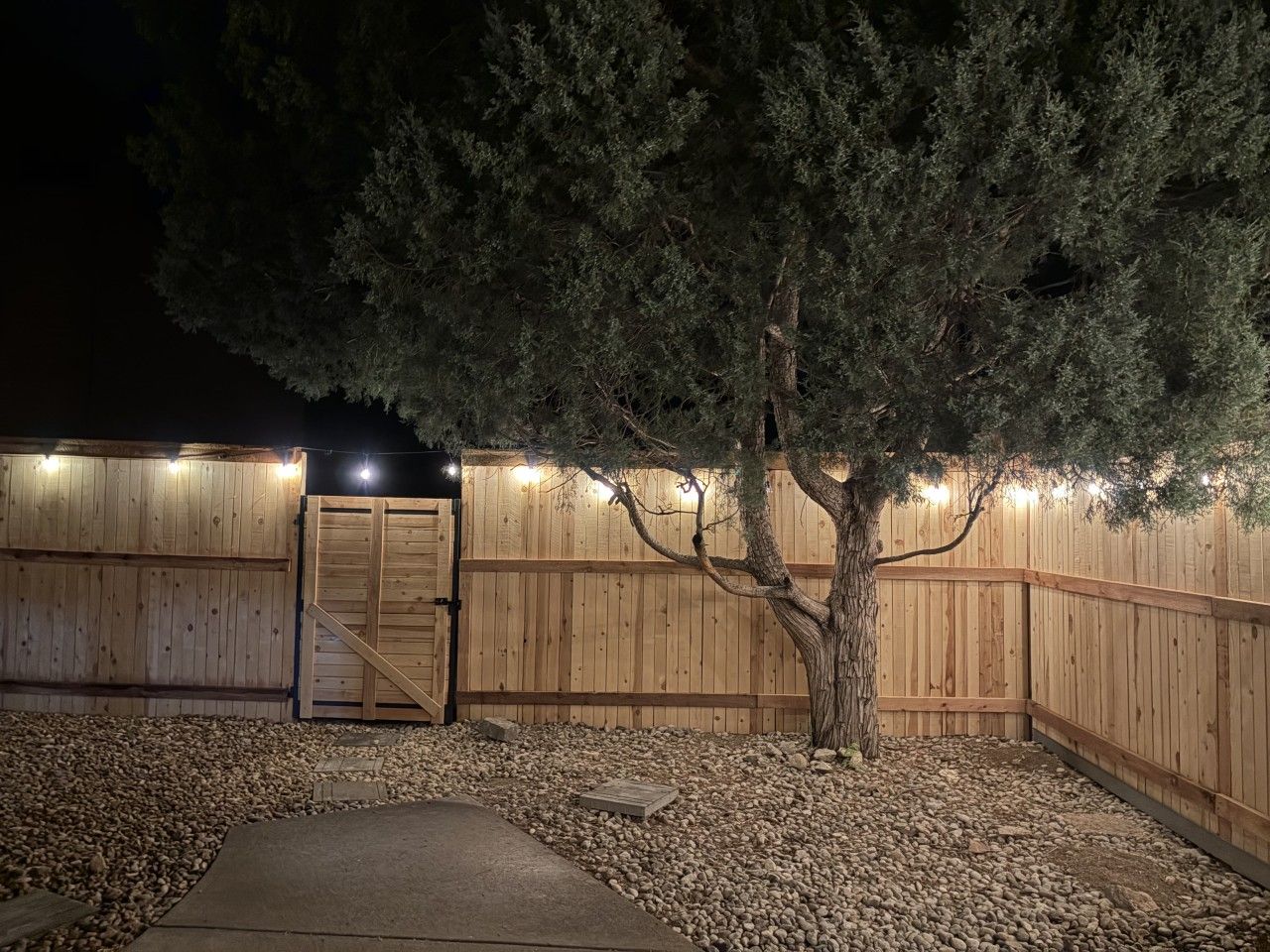 A wooden fence with a gated entrance, a tree, and a gravel pathway leading to the gate.