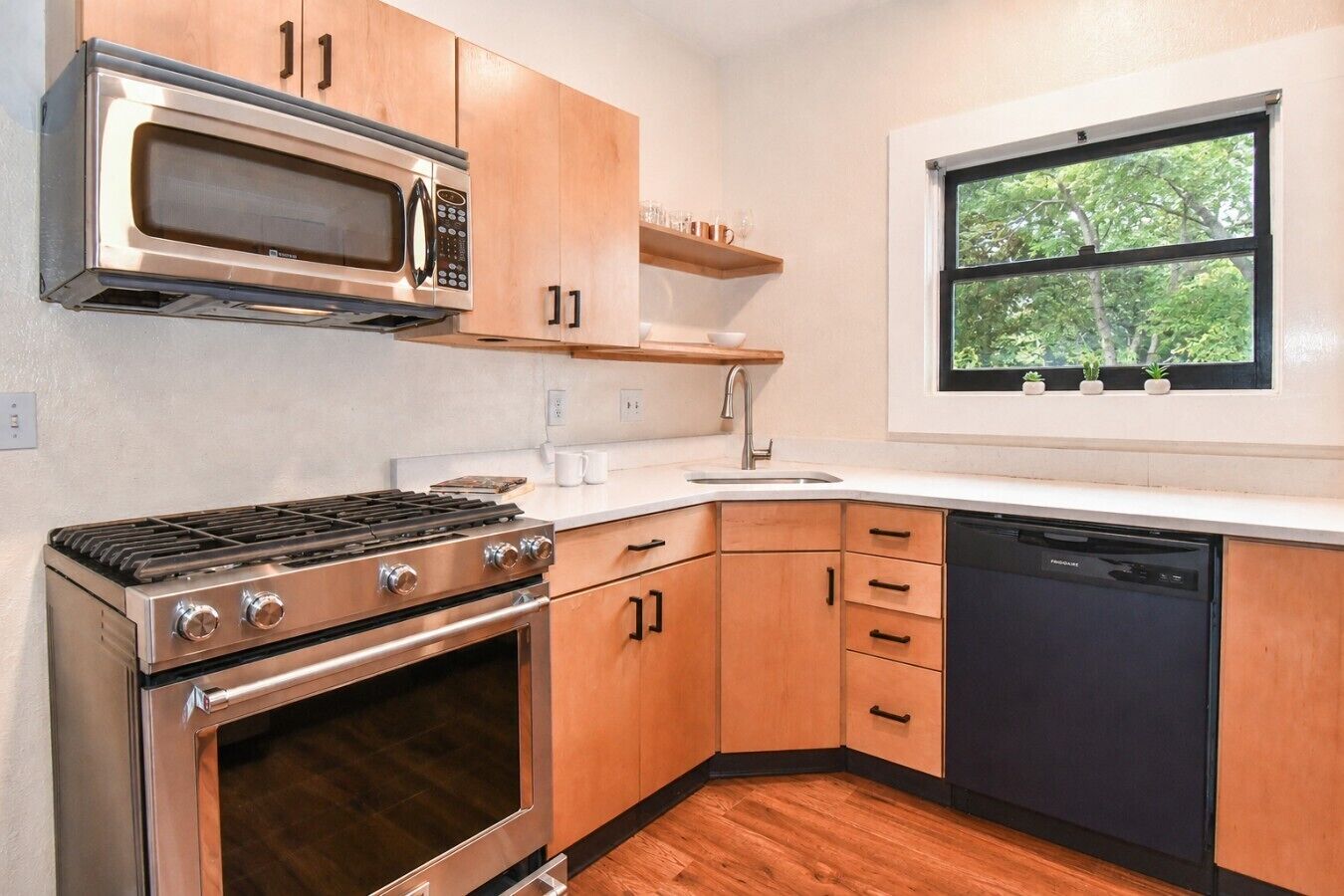 A modern kitchen with stainless steel appliances, wooden cabinets, and a window with a view of greenery.