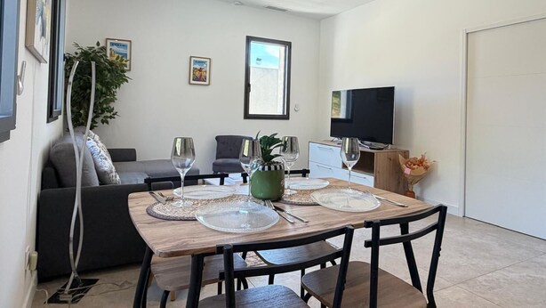 A dining area with a wooden table set for four, a grey sofa, a television, and a window with natural light.
