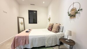 A bedroom with a bed, a bedside table with a lamp, a mirror, a window with blinds, and a wall decoration.
