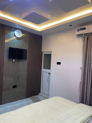 A modern bedroom with a bed, a flat-screen TV mounted on the wall, a ceiling light fixture, and an air conditioning unit. - The Haven | Modern 3BR En-suite Flat (Ibadan)