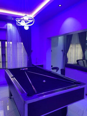 A pool table with a cue stick, a hanging light fixture, and a window with curtains. - The Haven | Modern 3BR En-suite Flat (Ibadan)