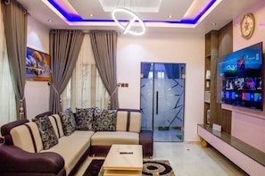 A modern living room with a sectional sofa, a flat-screen TV, and a glass door leading to a balcony. - The Haven | Modern 3BR En-suite Flat (Ibadan)