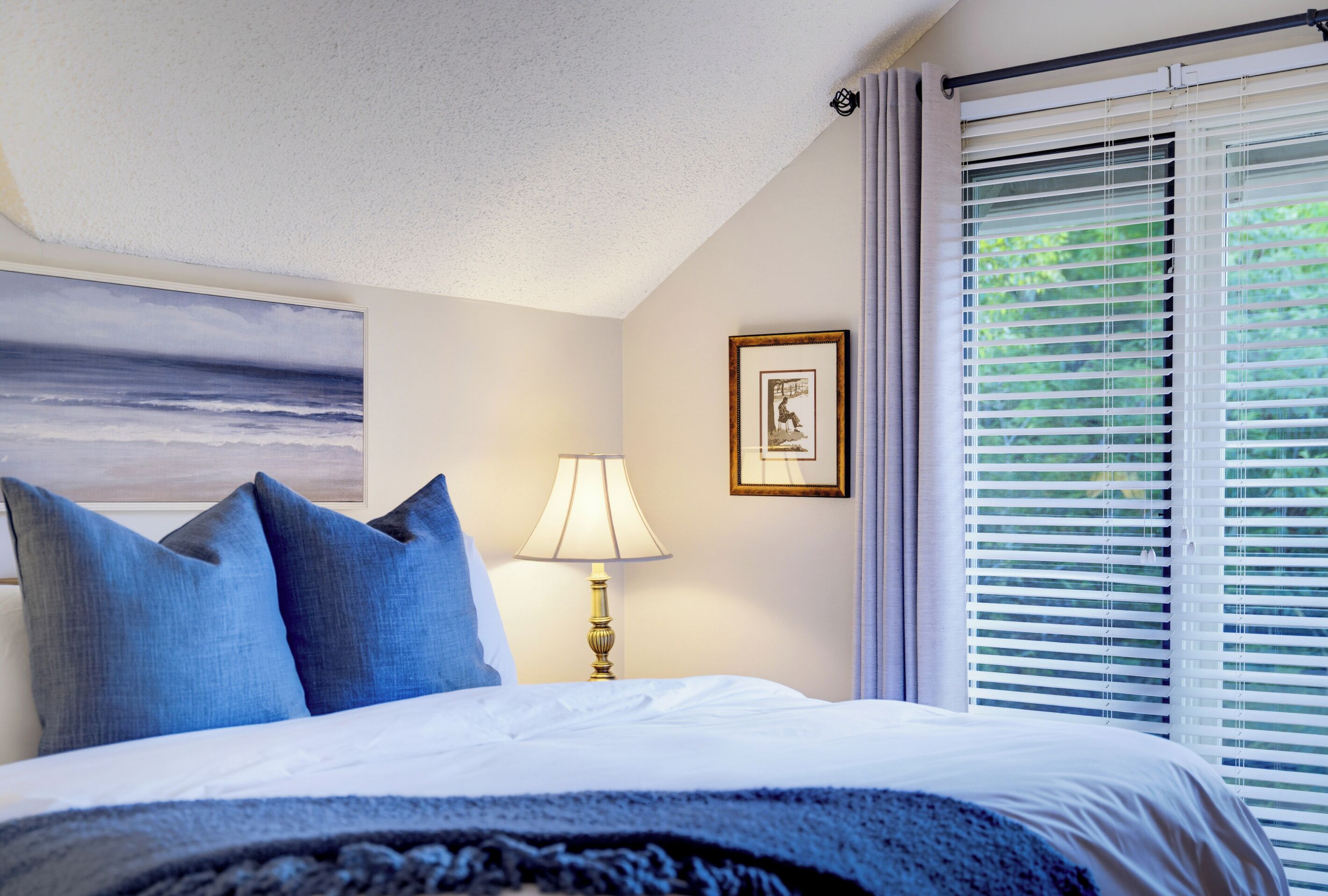A hotel room with a bed, a lamp, a framed picture, and a window with blinds.