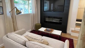 Una sala de estar con un sofá, una mesa de centro, un televisor montado en una pared negra y una ventana con cortinas.