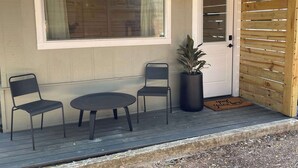 Una terraza de madera con una pequeña mesa, dos sillas, una planta en maceta y una puerta con un ventanal arriba.