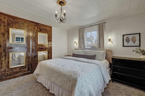 A bedroom with a large bed, wooden panel walls, a chandelier, and a window with curtains. - Pecan Grove Retreat - Pool & Fire pit (Fredericksburg)