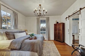 A bedroom with a bed, a chandelier, a wooden wardrobe, and a view of the outside through a window. - Pecan Grove Retreat - Pool & Fire pit (Fredericksburg)